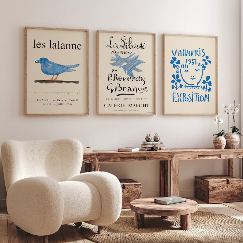 May include: Three framed art prints with a white background and blue text and illustrations. The left print features a blue bird with the text "les lalanne" and "CNAC 11, rue Berryer Paris B 5 juin-13 juillet 1975". The center print features a blue illustration of a bird with the text "La Libert&eacute; des mers par P.Reverdy G.Braqus. ET LIVRES ILLUSTRES RECENTS GALERIE MAEGHT". The right print features a blue illustration of a face with flowers around it and the text "VALAVRIS 1957 EXPOSITION".