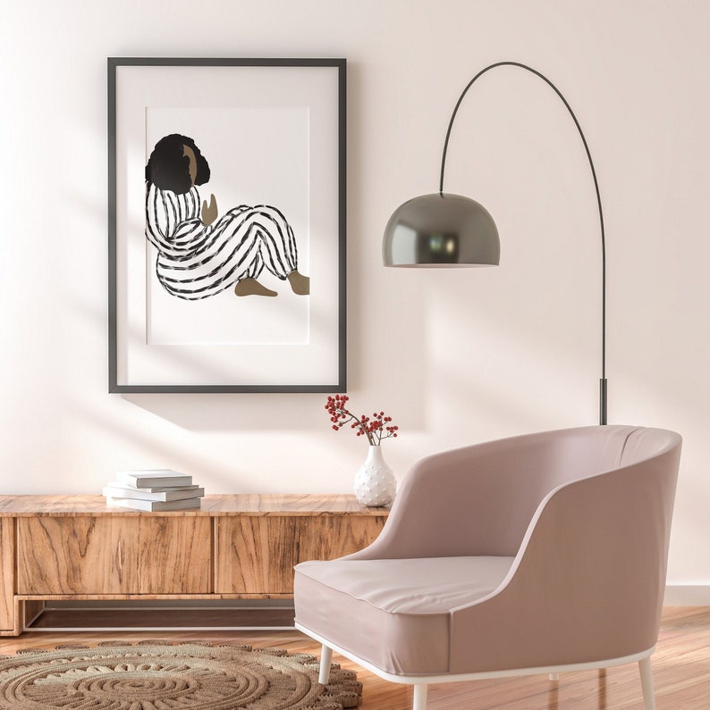 May include: A black and white illustration of a person in striped pajamas sitting on a white background. The illustration is framed in a black frame and is hanging on a white wall. A pink armchair sits in front of the artwork, and a vase with red berries sits on a wooden shelf.