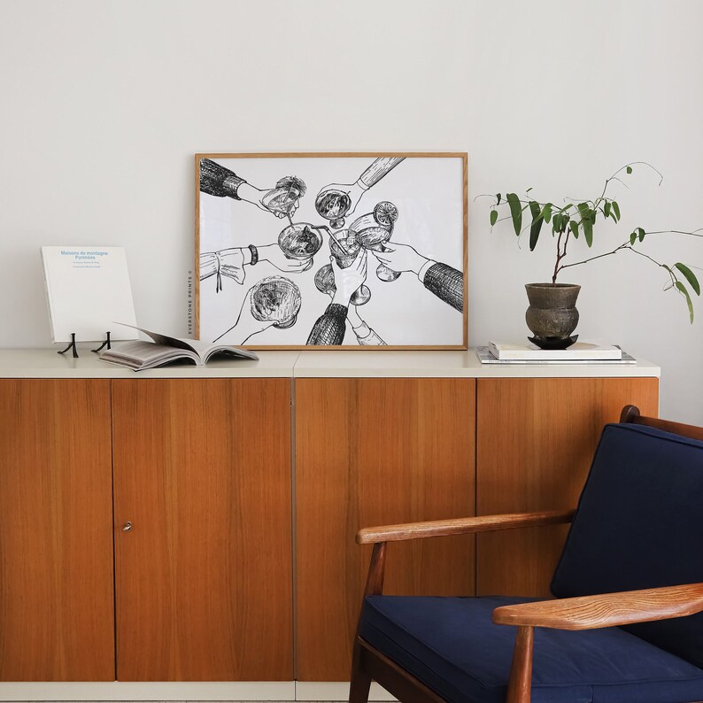 Op de afbeelding: Zwart-witte lijntekening van mensen die hun glazen heffen voor een toast. De tekening is ingelijst en hangt aan een muur boven een houten kast.