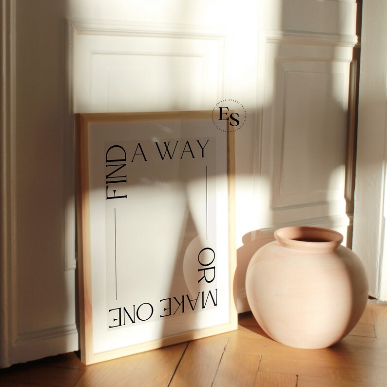 May include: A framed print with the text "FIND A WAY OR MAKE ONE" in black lettering on a white background. The print is leaning against a white wall next to a terracotta-colored vase.