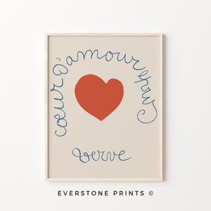 May include: A beige printable art print with a red heart and blue cursive text that reads "coeur d'amour" and "serve" in a circular shape around the heart. The print is framed in a light wood frame.