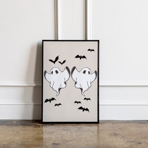 May include: A black framed print with two white ghosts and black bats on a beige background. The ghosts are facing each other with their arms outstretched. The bats are scattered around the ghosts.