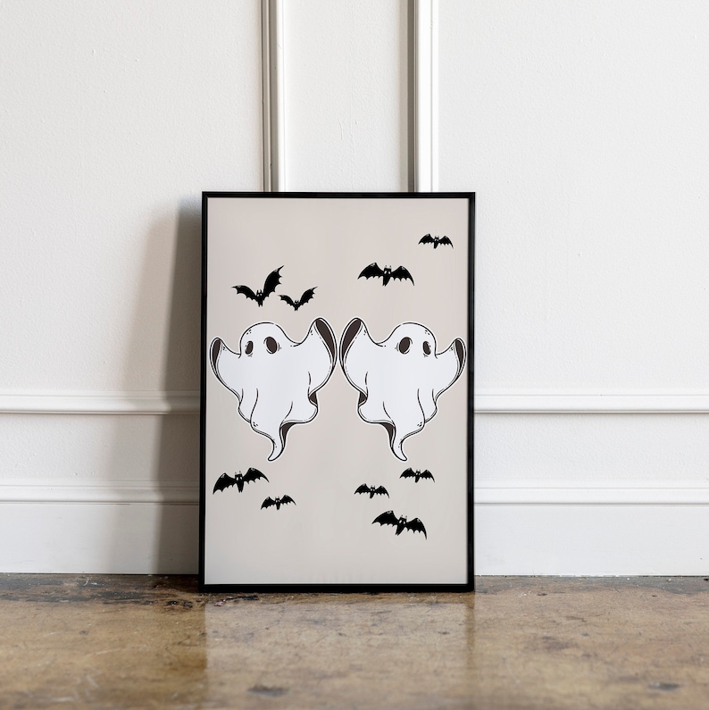May include: A black framed print with two white ghosts and black bats on a beige background. The ghosts are facing each other with their arms outstretched. The bats are scattered around the ghosts.