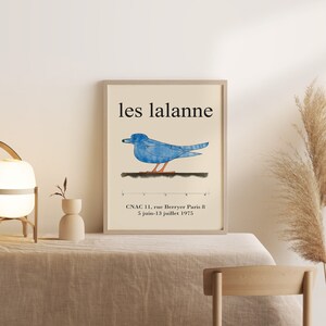 May include: A framed poster with the text "les lalanne" in black lettering. A blue bird with orange feet is depicted on a white background. The poster also includes the text "CNAC 11, rue Berryer Paris 8 5 juin-13 juillet 1975".