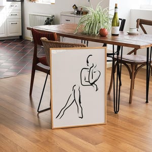 Pu&ograve; includere: Un disegno a linea nera e bianca incorniciato di una persona seduta con le gambe incrociate. Il disegno &egrave; su sfondo bianco ed &egrave; incorniciato in una cornice di legno chiaro. La cornice &egrave; appoggiata su un tavolo di legno con una pianta e una bottiglia di vino sullo sfondo.
