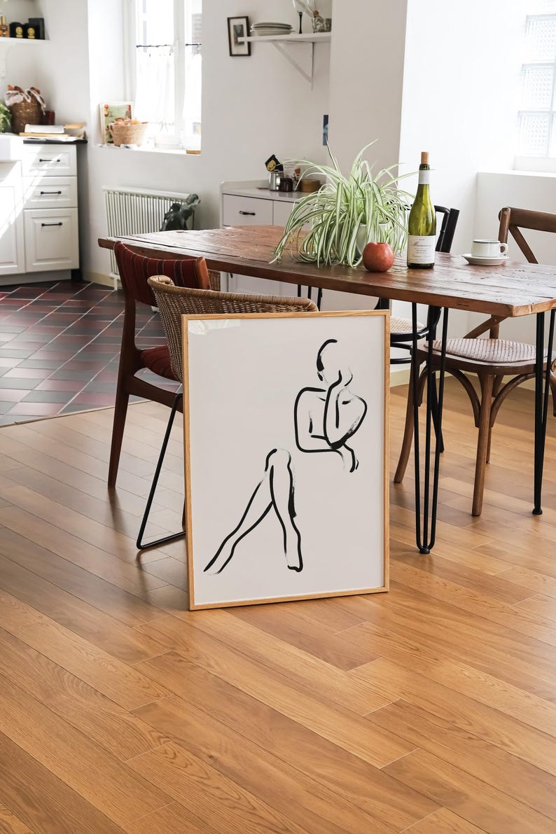 Pu&ograve; includere: Un disegno a linea nera e bianca incorniciato di una persona seduta con le gambe incrociate. Il disegno &egrave; su sfondo bianco ed &egrave; incorniciato in una cornice di legno chiaro. La cornice &egrave; appoggiata su un tavolo di legno con una pianta e una bottiglia di vino sullo sfondo.