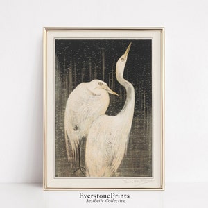 Op de afbeelding: Een vintage Japanse houtsnede met twee witte reigers die op een donkere, getextureerde achtergrond staan. De reigers staan ​​tegenover elkaar, met de nek van de ene reiger gebogen naar achteren. De print is onderaan rechts gesigneerd met "Hiroshi Yoshida".