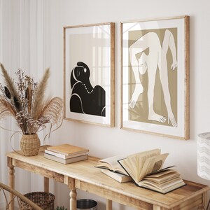 May include: Two framed minimalist line art prints of figures in black and white on a light brown background. The prints are hanging on a white wall above a wooden table with books.