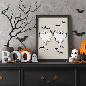 May include: A black framed picture with a white background and two white ghosts with black eyes and mouths. The ghosts are flying with their arms outstretched. There are black bats flying around the ghosts. The picture is on a black dresser with a BOO sign and pumpkins.