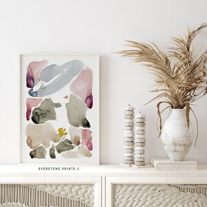 May include: An abstract watercolour painting of a woman wearing a hat, featuring shades of pink, blue, brown, and green. The painting is framed and displayed on a white shelf with a vase of dried pampas grass and decorative candle holders.