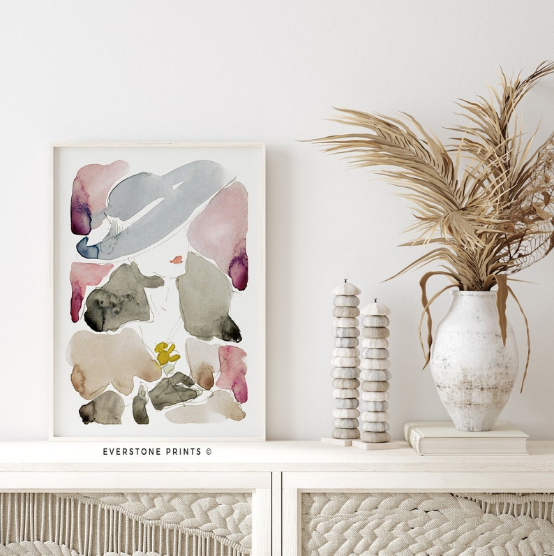 May include: An abstract watercolour painting of a woman wearing a hat, featuring shades of pink, blue, brown, and green. The painting is framed and displayed on a white shelf with a vase of dried pampas grass and decorative candle holders.