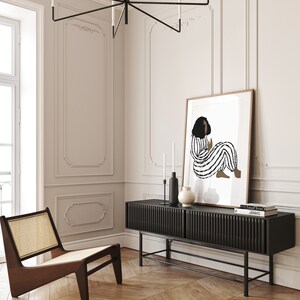 May include: A black and white illustration of a person in a striped outfit sitting on a white background. The illustration is framed and hanging on a white wall above a black media console with a wood top. The console has a black metal frame and is sitting on a wood floor.