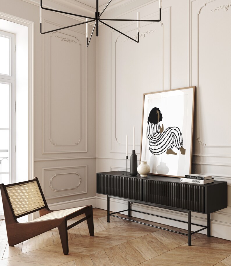 May include: A black and white illustration of a person in a striped outfit sitting on a white background. The illustration is framed and hanging on a white wall above a black media console with a wood top. The console has a black metal frame and is sitting on a wood floor.