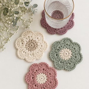 Könnte beinhalten: Vier handgefertigte gehäkelte Blumenuntersetzer in Creme-, Mauve- und Grüntönen. Ein klares Glas mit Goldrand steht auf einem Untersetzer. Die Untersetzer liegen auf einer weißen Oberfläche, mit einem Zweig weißer Blumen im Hintergrund.
