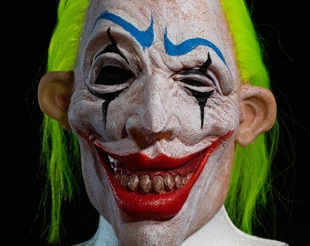 May include: A creepy clown mask with a wide, toothy grin. The mask is white with blue and black painted details. The clown has bright yellow hair.