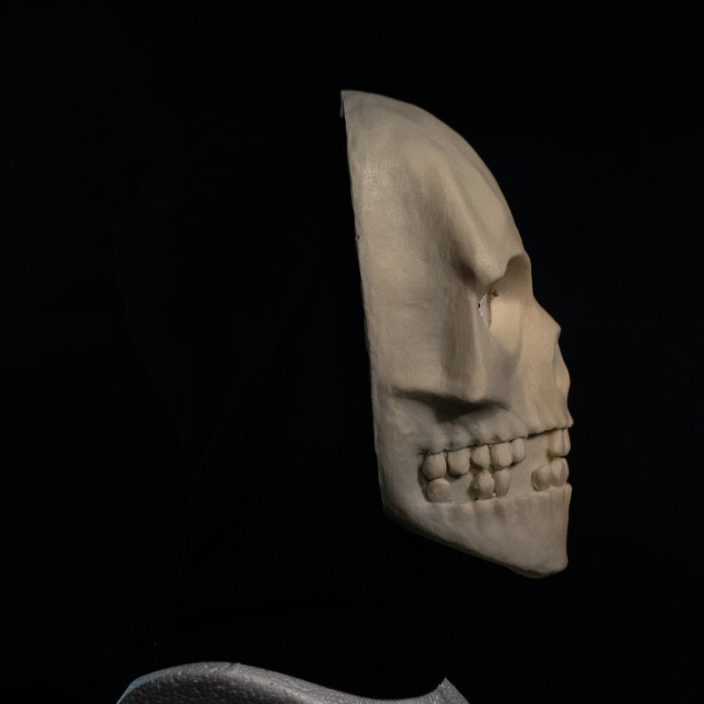 May include: A white skull mask with a black hood covering the back of the head. The skull is shown in profile, with the right side of the face visible. The mask has a detailed sculpt of the teeth and eye sockets.