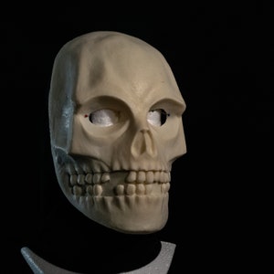 May include: A white skull mask with detailed features, including teeth and eye sockets. The mask is made of a smooth, textured material and is displayed on a black stand.