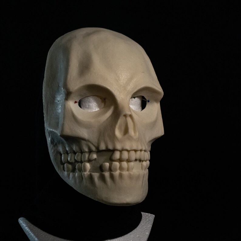 May include: A white skull mask with detailed features, including teeth and eye sockets. The mask is made of a smooth, textured material and is displayed on a black stand.