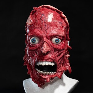 May include: A highly detailed Halloween mask showing a peeled-back face with exposed muscle and tissue. The mask has bright blue eyes, a wide open mouth, and intricate textures, creating a terrifying effect. The mask is mainly red with areas of exposed skin.