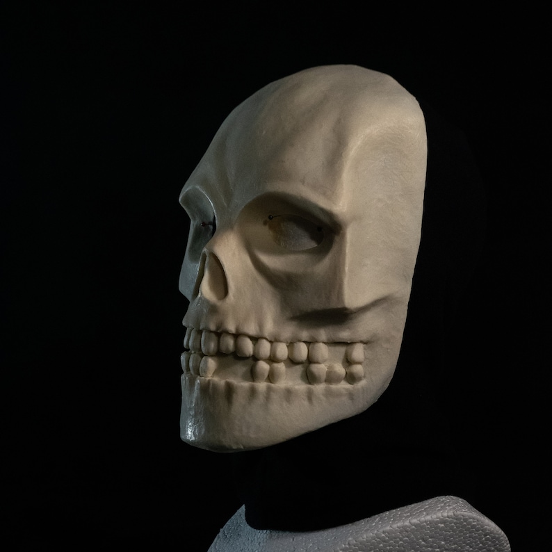 May include: A white skull mask with a detailed, realistic design. The mask has a closed mouth with teeth and a single eye socket.