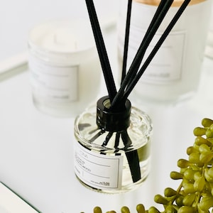 May include: A glass diffuser bottle with black reeds, a white label, and a black cap. Two white candles in glass jars are in the background. A green plant is in the foreground. The diffuser is labelled "SONA".