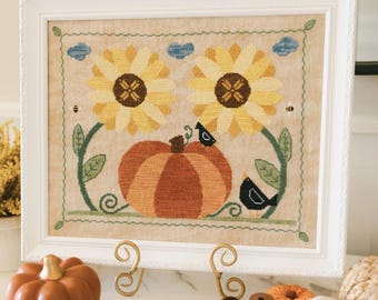 Puede incluir: Una bordadura de punto de cruz enmarcada con dos girasoles amarillos, una calabaza y dos pájaros negros. La bordadura está hecha en una tela beige con un borde marrón. El marco es blanco con un borde decorativo.