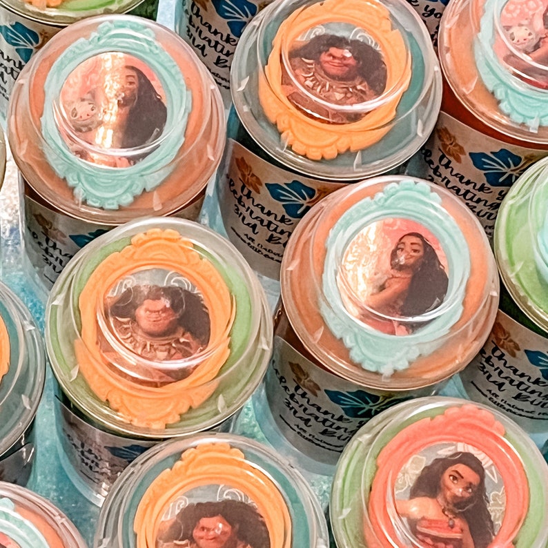 K&ouml;nnte beinhalten: Eine Sammlung durchsichtiger Plastikbeh&auml;lter mit bunten Deckeln, die mit Bildern von Figuren aus dem Animationsfilm Vaiana verziert sind. Die Deckel haben ein mehrschichtiges Design mit orangefarbenen, gr&uuml;nen und blauen Akzenten. Die Beh&auml;lter sind mit den Worten "Vielen Dank, dass Sie mit uns feiern!" beschriftet.