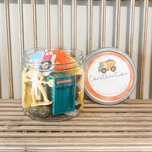 May include: A clear glass jar filled with various construction-themed toys, including a yellow construction worker figurine, a green construction sign, and a red toy truck. The jar has a silver lid with a label that reads "Little Fox and Wren Co. Construction,  Cause + Effect, All Natural Play Dough."