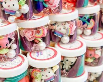 Puede incluir: Frascos transparentes llenos de juguetes coloridos, incluyendo unicornios, arcoíris y flores. Cada frasco tiene una tapa blanca con un borde rojo. Algunas tapas tienen el texto "Thank You". Los frascos están apilados.