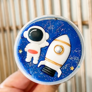 May include: A blue slime with a white astronaut and a white rocket ship with gold accents. The slime is in a clear plastic container.