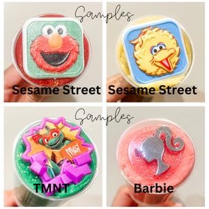 K&ouml;nnte beinhalten: Vier durchsichtige Plastikbecher mit buntem Schleim gef&uuml;llt. Jeder Becher hat einen anderen Charakter-Topper: Elmo, Big Bird, Teenage Mutant Ninja Turtles und Barbie. Die Becher sind mit "Sesame Street", "Sesame Street", "TMNT" und "Barbie" beschriftet.