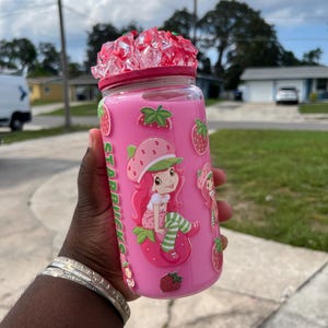 May include: A pink Starbucks tumbler featuring a Strawberry Shortcake design. The tumbler has a pink lid with clear and pink crystals. The design includes the Starbucks logo and images of Strawberry Shortcake and strawberries.