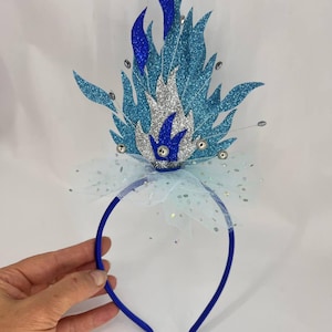 Può includere: Una fascia blu con un design a fiamma decorativa. La fiamma è realizzata con materiali blu e argento glitterati, con accenti di gemme trasparenti. Un fiocco di tulle bianco si trova alla base della fiamma.