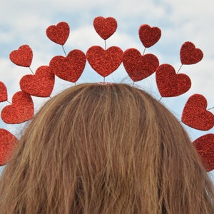 May include: A red heart-shaped headband with a row of red glitter hearts. The headband is worn on a person's head.