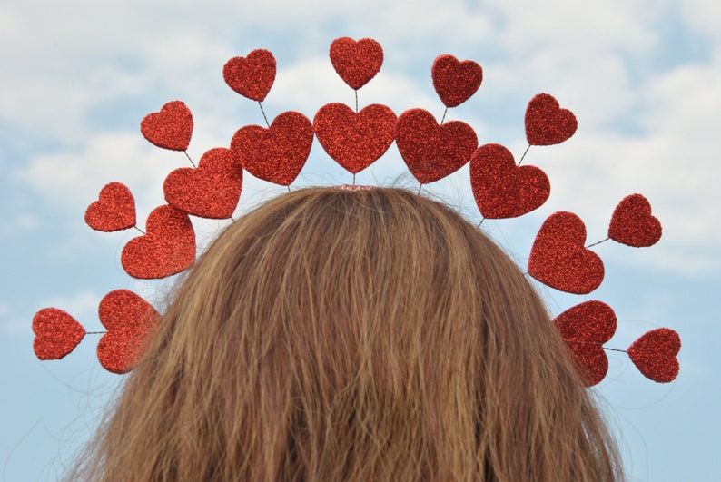 May include: A red heart-shaped headband with a row of red glitter hearts. The headband is worn on a person's head.