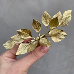 May include: A gold-colored hair comb featuring a leaf design. The comb has multiple leaves attached to a central stem, with fine details and texture on each leaf. The leaves are arranged in a symmetrical pattern, creating an elegant accessory.