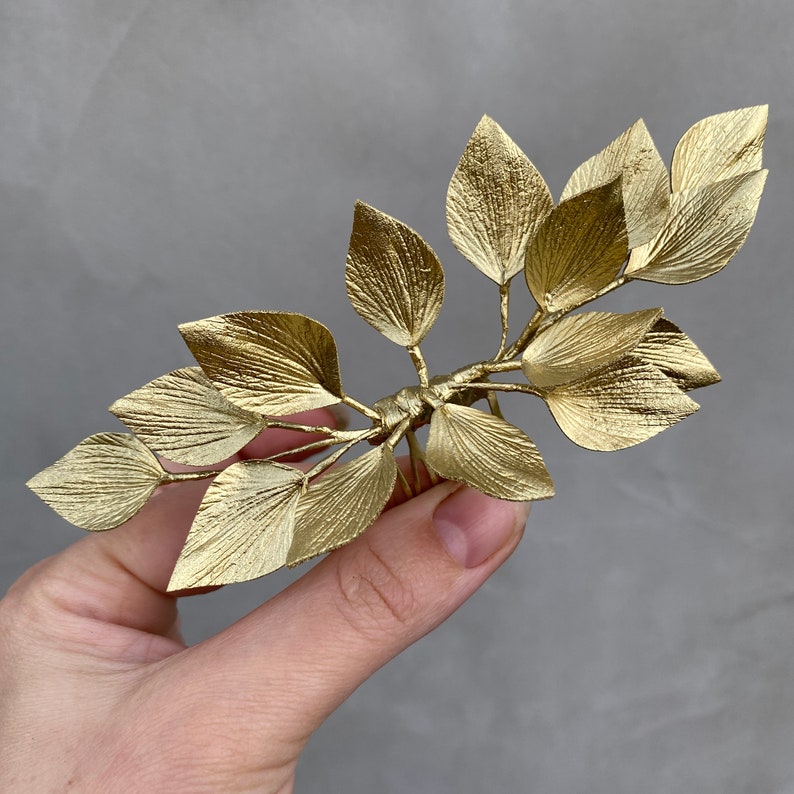 May include: A gold-colored hair comb featuring a leaf design. The comb has multiple leaves attached to a central stem, with fine details and texture on each leaf. The leaves are arranged in a symmetrical pattern, creating an elegant accessory.