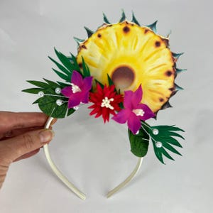 May include: A white headband with a pineapple, green leaves, and pink and red flowers. The headband is decorated with white pearl beads.
