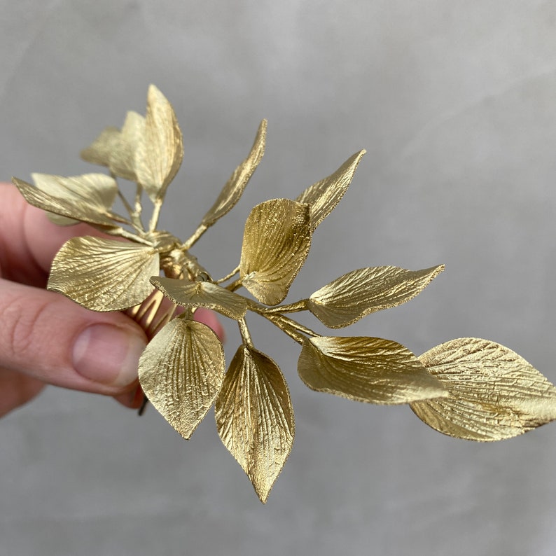 May include: A gold-colored leaf hair accessory. The leaves have detailed veining and are arranged in a delicate, naturalistic design. The accessory is held in a hand against a neutral background, showcasing its metallic sheen and intricate details.