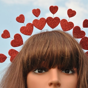 May include: A red glitter heart headband with a row of hearts around the top. The headband is worn on a mannequin with brown hair.