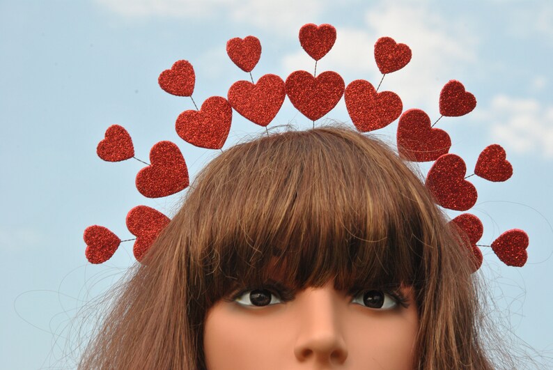 May include: A red glitter heart headband with a row of hearts around the top. The headband is worn on a mannequin with brown hair.