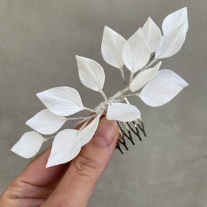 May include: A delicate white leaf hair comb. The comb features multiple white leaves attached to a silver-toned wire, with a gold-colored comb base. The leaves have detailed veining, creating a realistic appearance. Ideal for weddings or special occasions.