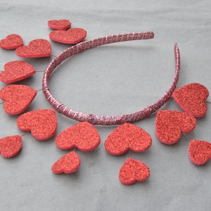 May include: A silver headband with red glitter hearts attached. The hearts are arranged in a circle around the headband, with some hanging down.