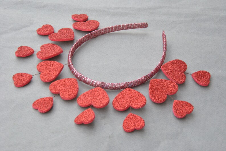 May include: A silver headband with red glitter hearts attached. The hearts are arranged in a circle around the headband, with some hanging down.