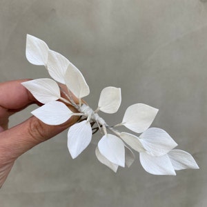 May include: A delicate, white floral hair accessory featuring multiple paper-like leaves. The leaves are clustered together and attached to a thin, flexible stem. The accessory is held against a neutral background.