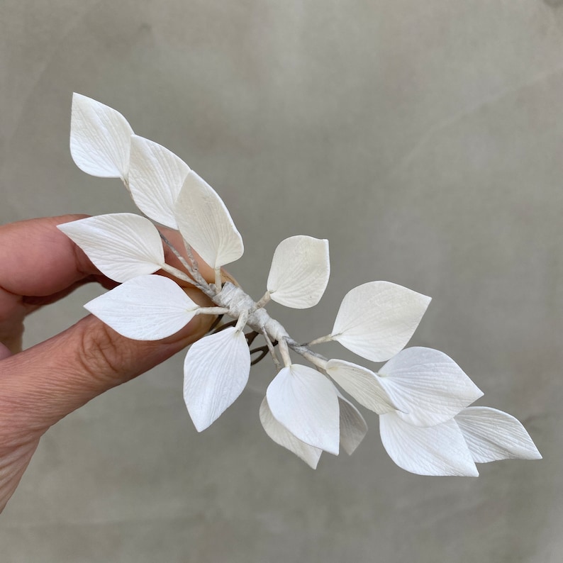 May include: A delicate, white floral hair accessory featuring multiple paper-like leaves. The leaves are clustered together and attached to a thin, flexible stem. The accessory is held against a neutral background.