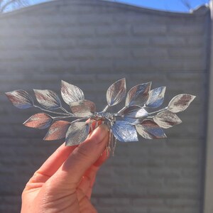 May include: A silver-toned hair comb featuring a leaf design. The leaves have a metallic finish with shades of silver and copper. The comb is held in a hand against a blurred background.
