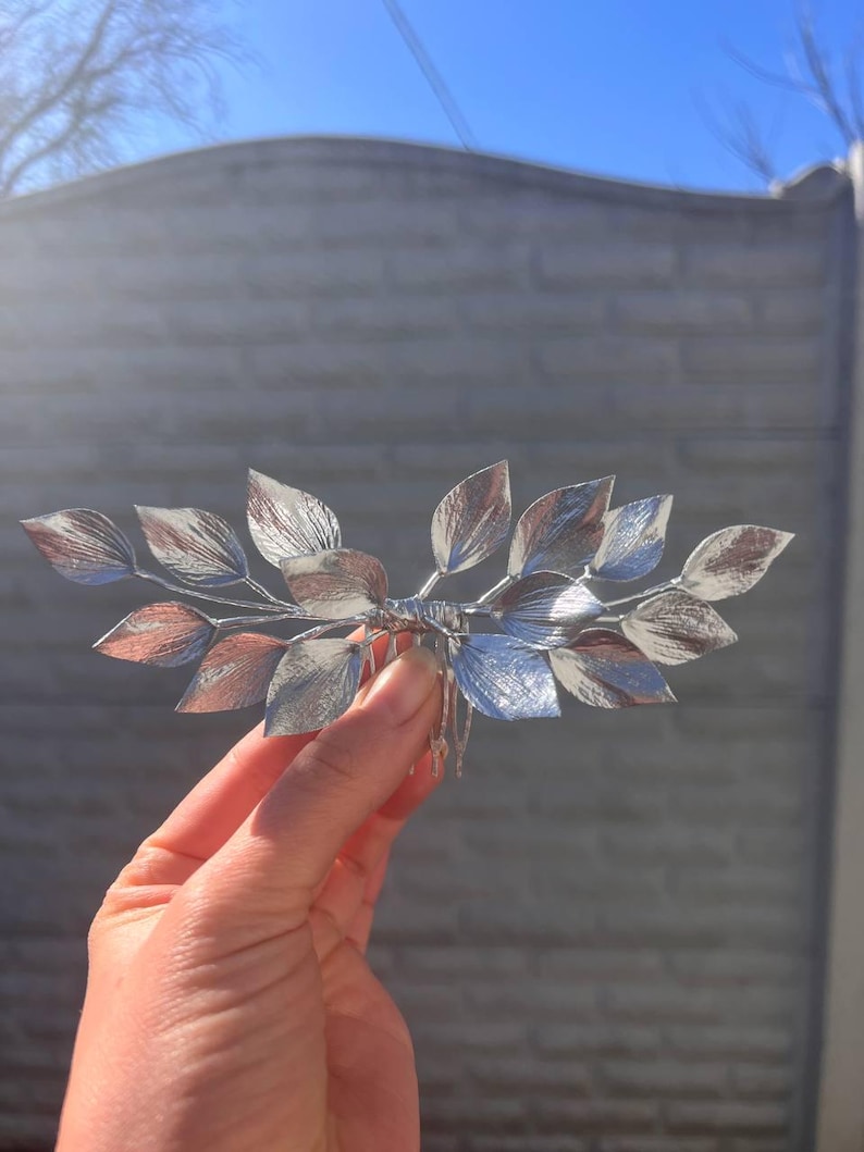 May include: A silver-toned hair comb featuring a leaf design. The leaves have a metallic finish with shades of silver and copper. The comb is held in a hand against a blurred background.
