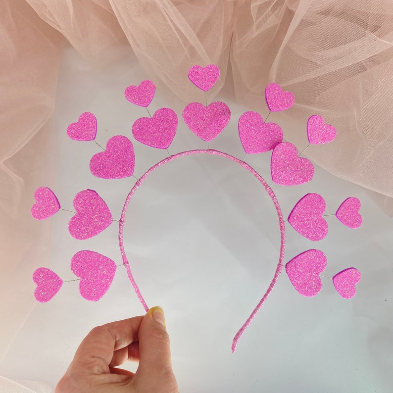 May include: Pink headband with a row of pink glitter hearts attached to it. The headband is made of a thin, flexible material.