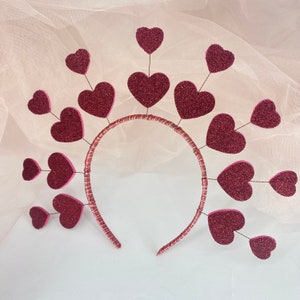 May include: A pink headband with a circle of pink hearts attached to it. The hearts are all different sizes and are arranged in a circle around the headband.